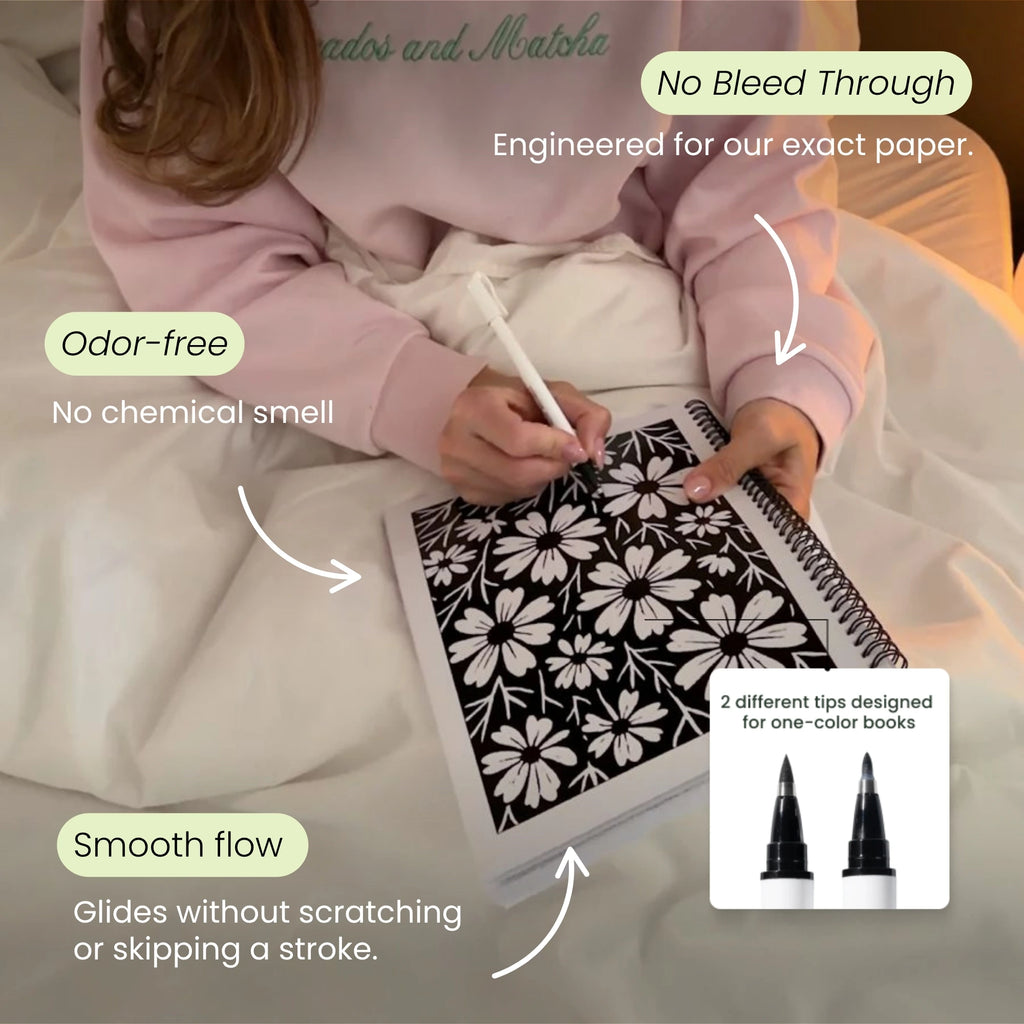 Person in a pink jumper colouring a black floral page in bed, with no-bleed and smooth-flow callouts