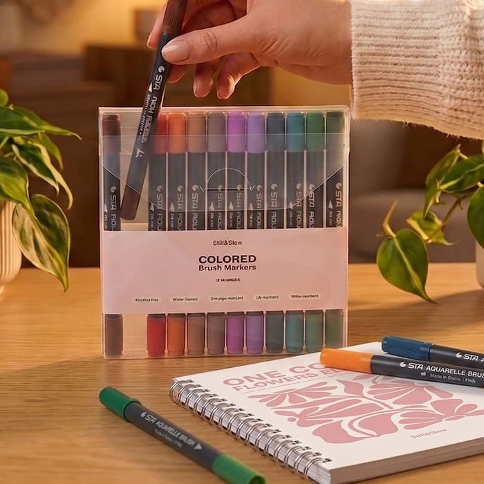 Hand lifting a marker from the Colored Brush Markers box on a table, next to a pink spiral coloring book