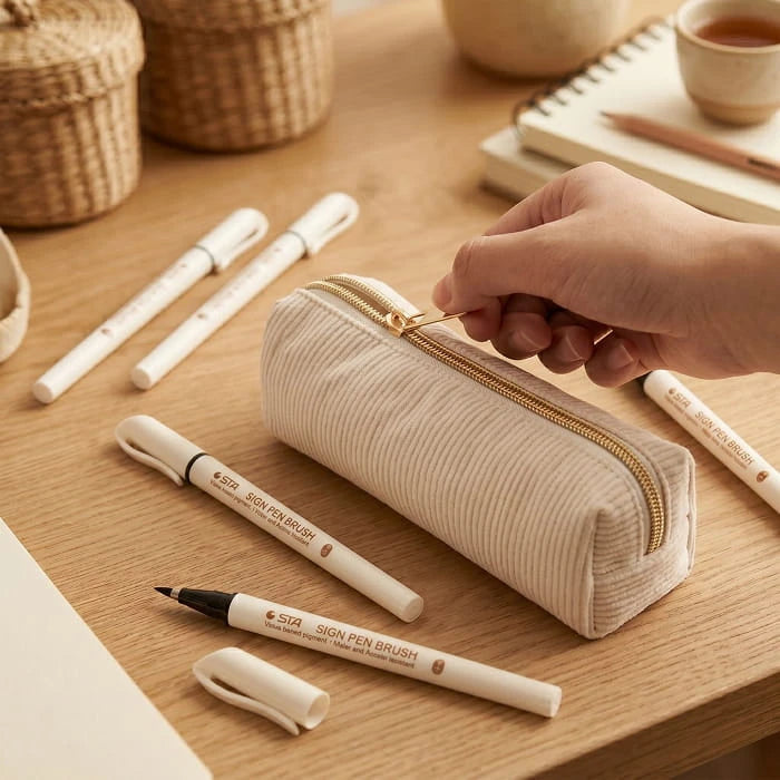 Hand opening the beige corduroy pencil case on a wooden table beside four white brush pens