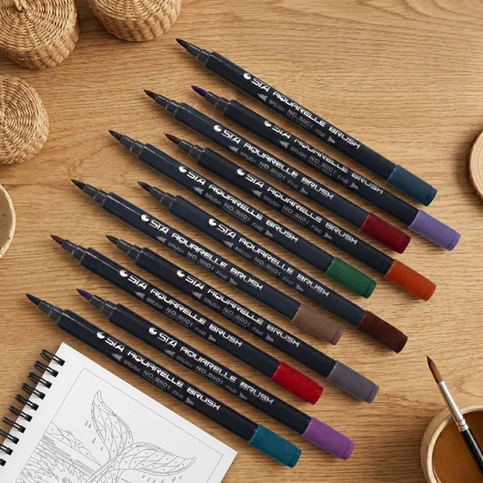 Twelve coloured brush markers arranged on a wooden table beside an open coloring book